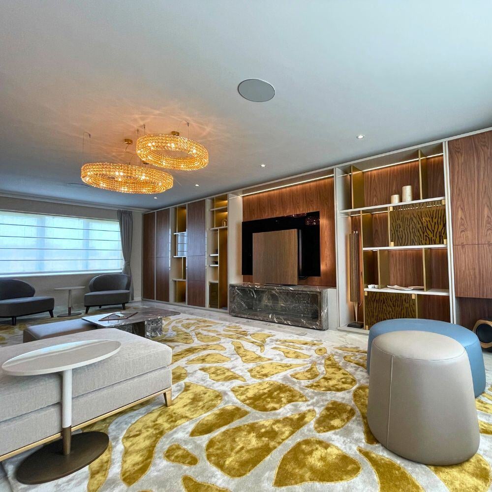 Luxurious media room featuring golden chandeliers, custom wooden shelving, and plush gold-accented carpet over marble flooring.